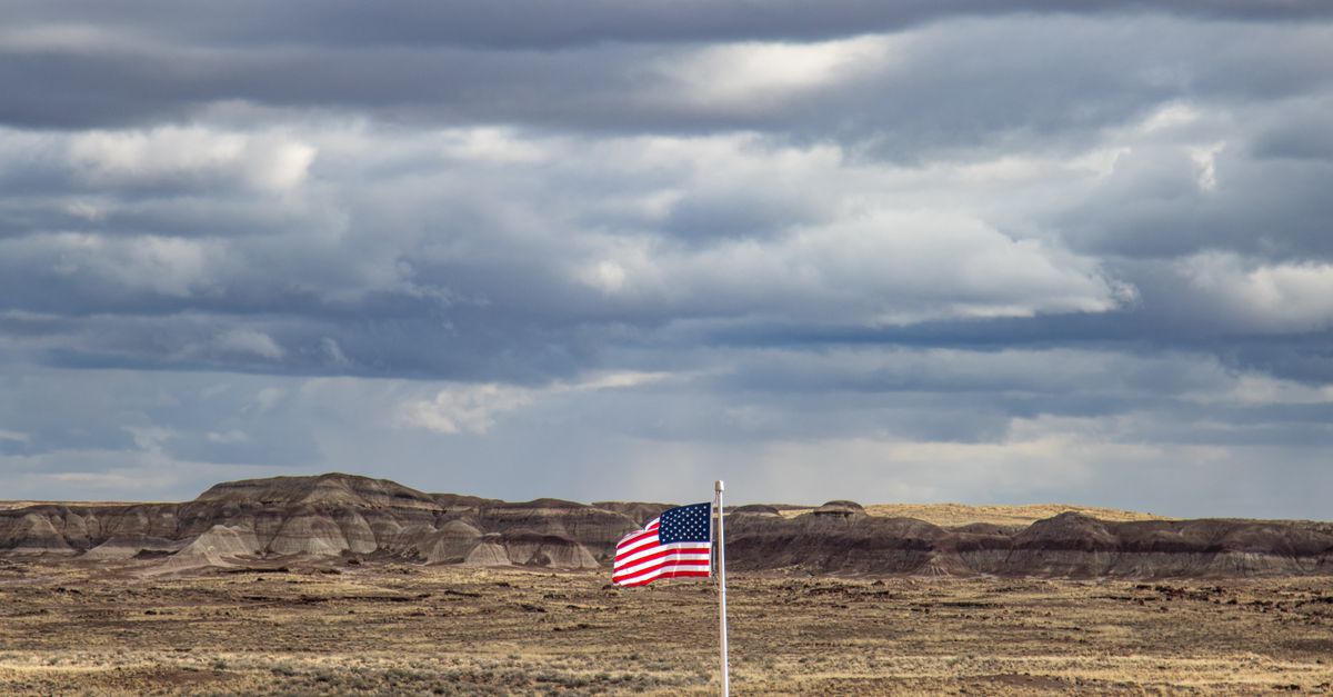 An American flag stands tall in a vast desert landscape under a dramatic cloudy sky.